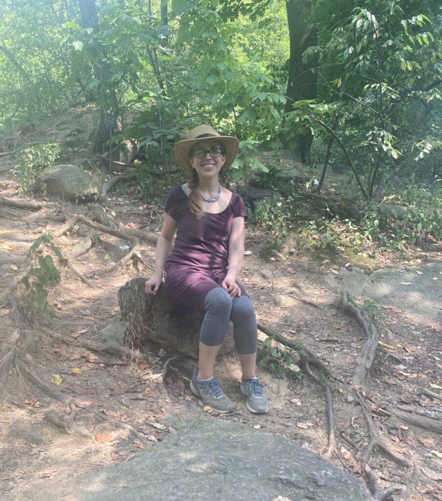 Dr. Biehler, a white woman in a purple dress and sun hat, smiling at camera while sitting on a rock surrounded by trees